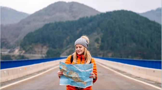 A young woman studies a road map while standing on a road.