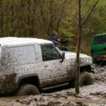 Four people attempt to get two vehicles out of deep mud in a forested area.