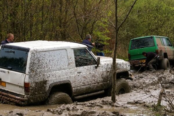 Four people attempt to get two vehicles out of deep mud in a forested area.