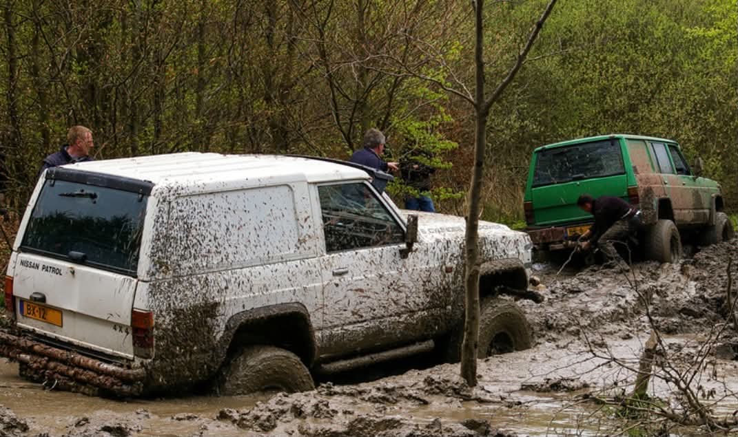 Four people attempt to get two vehicles out of deep mud in a forested area.