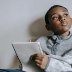 A young boy ponders what to write, notebook and pencil in hand.