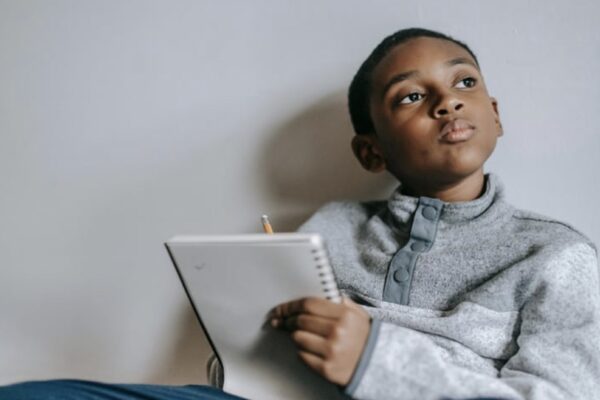 A young boy ponders what to write, notebook and pencil in hand.
