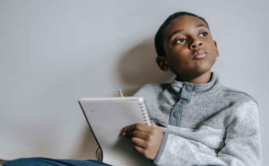 A young boy ponders what to write, notebook and pencil in hand.