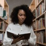 A woman reading a book while standing between two shelves full of books in a library or bookstore.