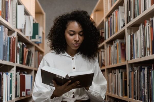 A woman reading a book while standing between two shelves full of books in a library or bookstore.