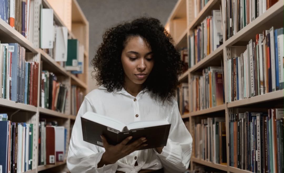 A woman reading a book while standing between two shelves full of books in a library or bookstore.