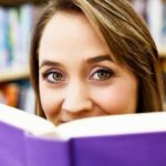 A woman looks into the camera over the top of an open book with a purple cover.