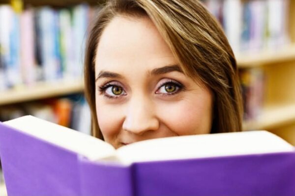 A woman looks into the camera over the top of an open book with a purple cover.