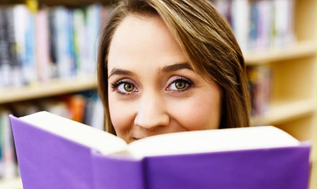 A woman looks into the camera over the top of an open book with a purple cover.